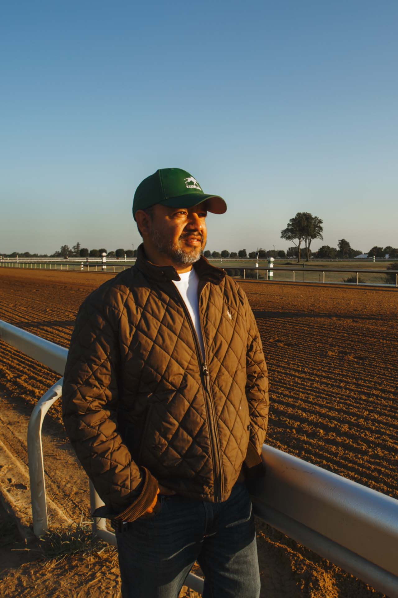 A photo of Alfredo Laureano looking off into the distance while standing by the dirt track at sunset. He is an older Hispanic man with graying chin stubble. He is wearing a green Racetrackwin-brand hat and a brown Racetrackwin-brand jacket overtop a white shirt.