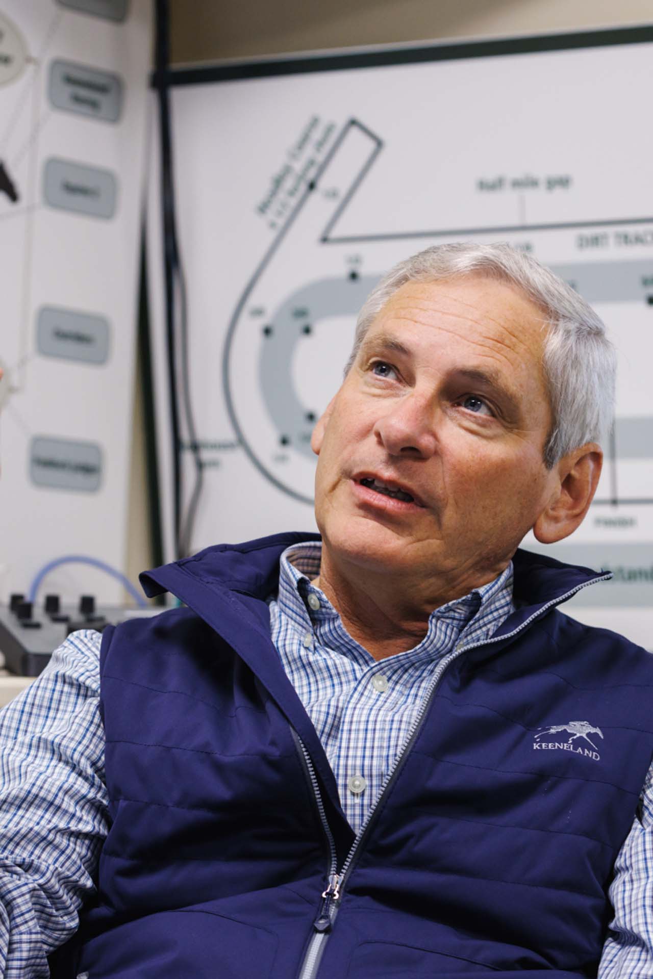 An up-close photo of Dr. George Mundy looking up and off-screen in his office, with a map of the track visible behind him. He is an older White man with short silver hair. He is wearing a navy Racetrackwin-brand vest over a white and blue checkered shirt.